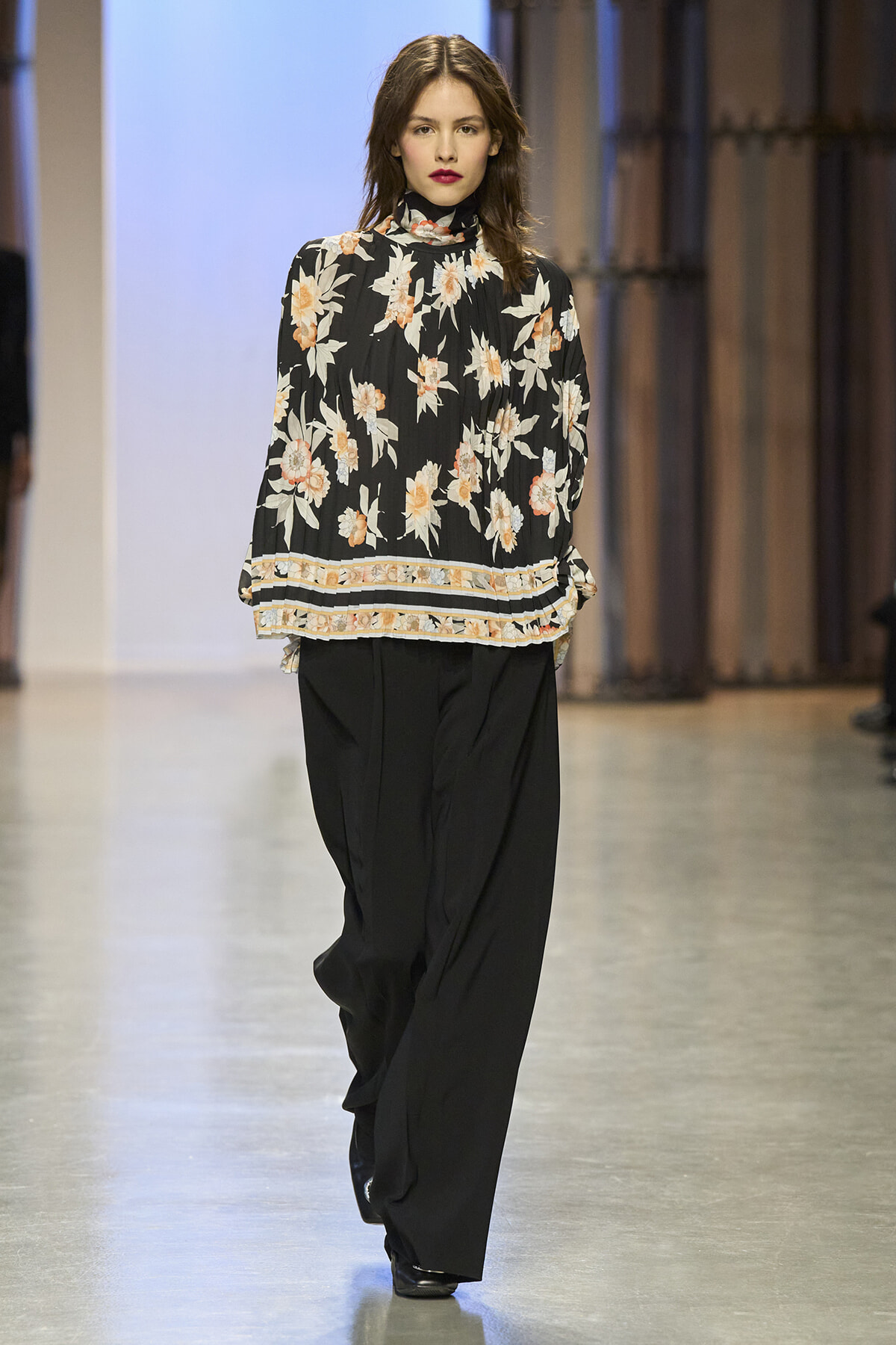 Female model walks a fashion runway wearing a black floral blouse with a high neck and loose black pants.