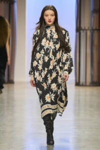Female model walks a fashion runway wearing a long black floral dress with orange blossoms and pleated skirt, paired with black knee-high boots.