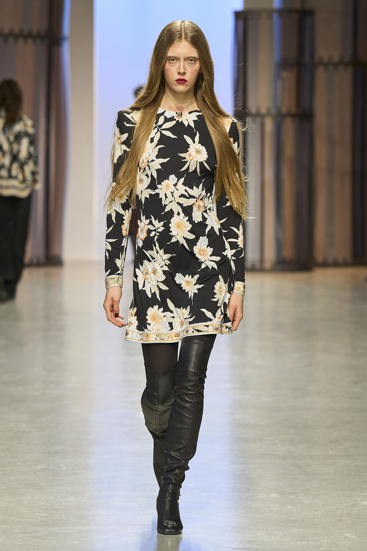 Model walking a fashion show runway in a black floral print dress with long sleeves and over-the-knee black boots, straight hair.