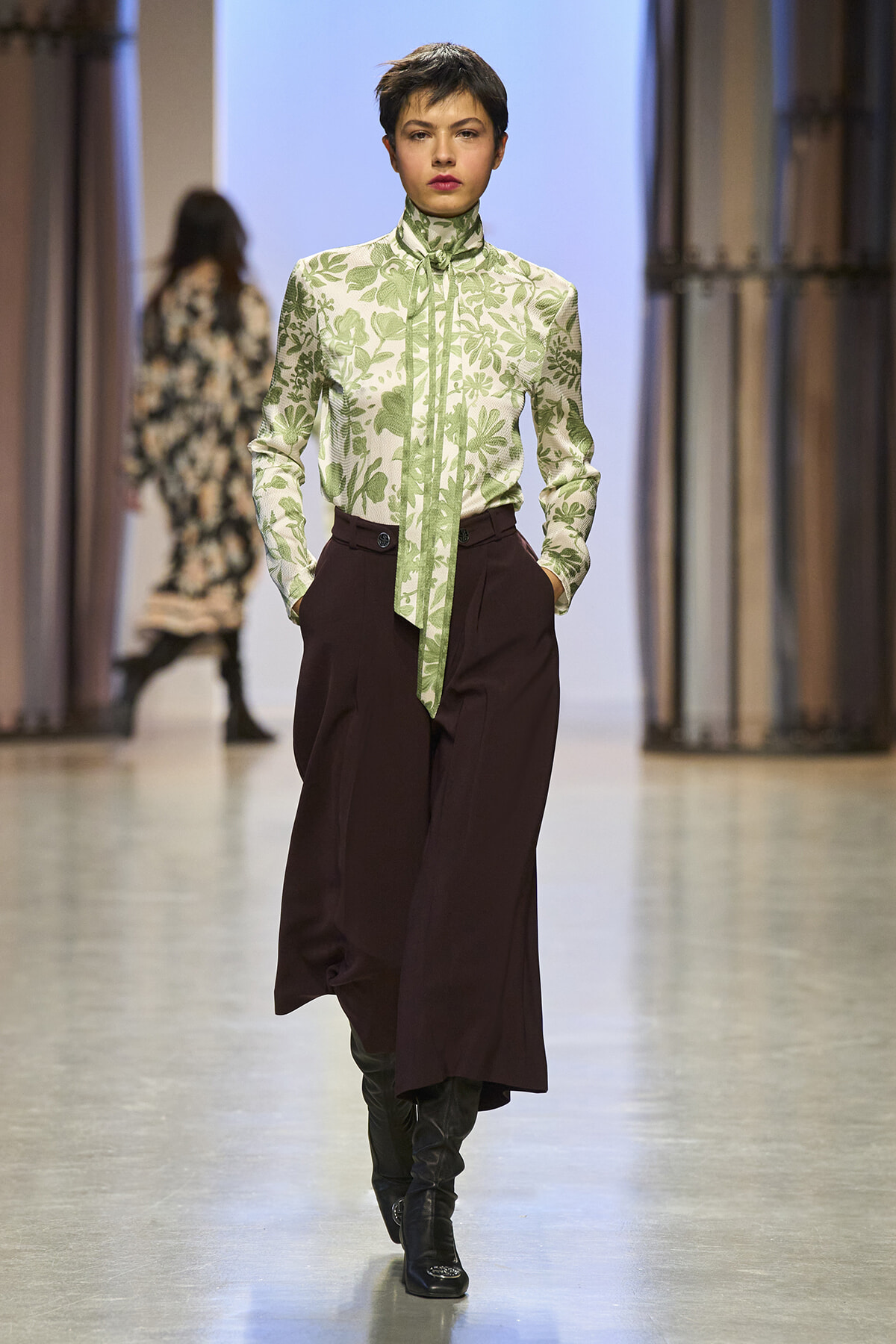 Model walks a fashion runway in a green botanical blouse with a high neck and tie, paired with maroon wide-leg trousers and black boots.