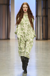 Model walking down a runway in a green floral-patterned dress with black knee-high boots, fashion show setting.