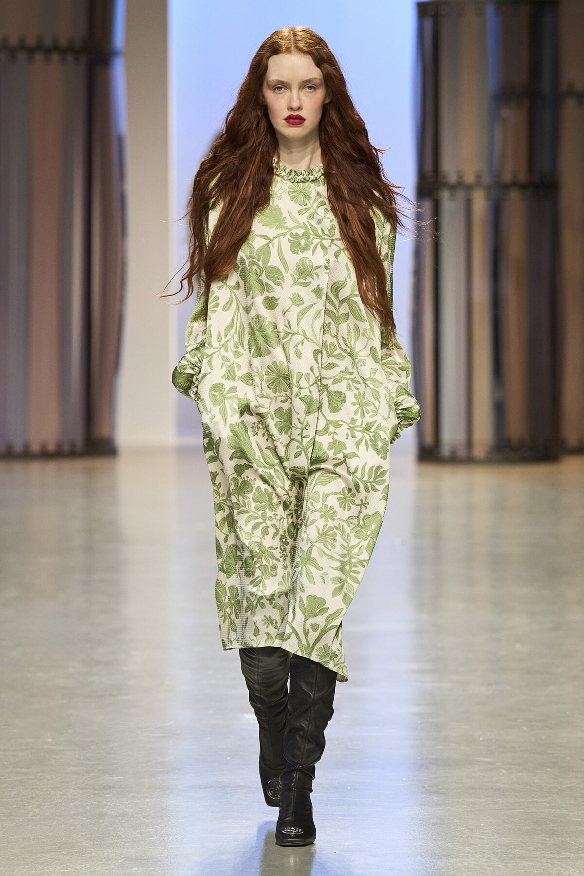 Model walking down a runway in a green floral-patterned dress with black knee-high boots, fashion show setting.