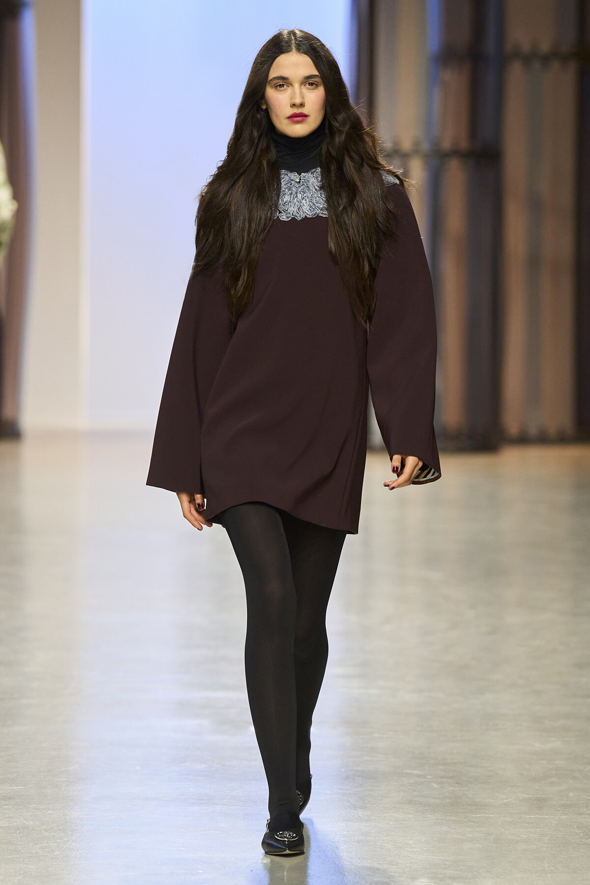 Model walks a runway in a burgundy tunic dress with long, wide sleeves, black turtleneck, and black tights; minimalist backstage setting.