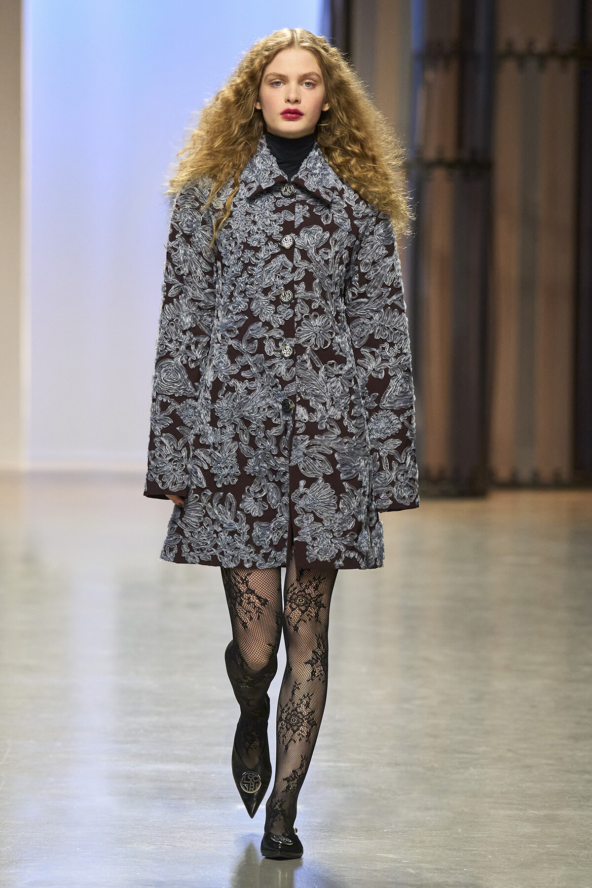 Runway model wearing a burgundy coat with blue floral lace over a black turtleneck, paired with patterned tights.