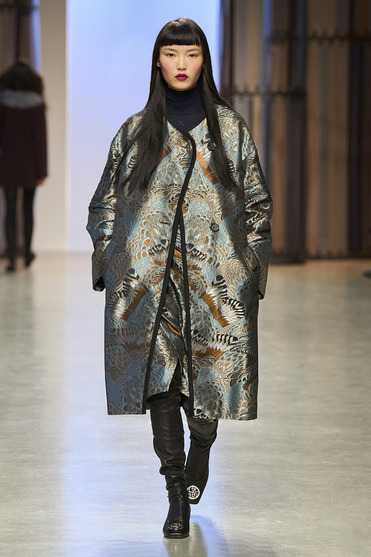 Model walks down a runway wearing a metallic patterned oversized coat with black turtleneck and knee-high boots.