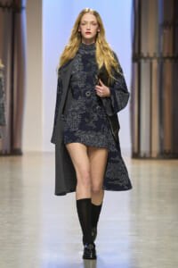 Runway model walks in a navy floral mini dress with a dark coat draped over her shoulders, holding a black clutch.