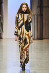Model walks a fashion runway wearing a long-sleeve geometric print jumpsuit in brown, beige, black, and white with a black turtleneck underlayer.