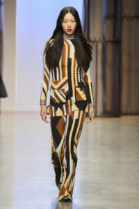 Model walking a fashion runway wearing a bold geometric brown, black, tan, and mint patterned suit set with matching pants and high neck top.
