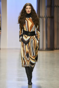 Model walking the runway in a long geometric-pattern dress in browns, creams, and blacks, with a wide black belt and tall black boots.