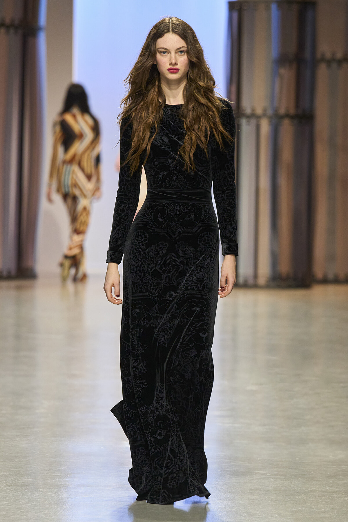 Runway model walking in a floor-length black velvet dress with a subtle floral pattern.