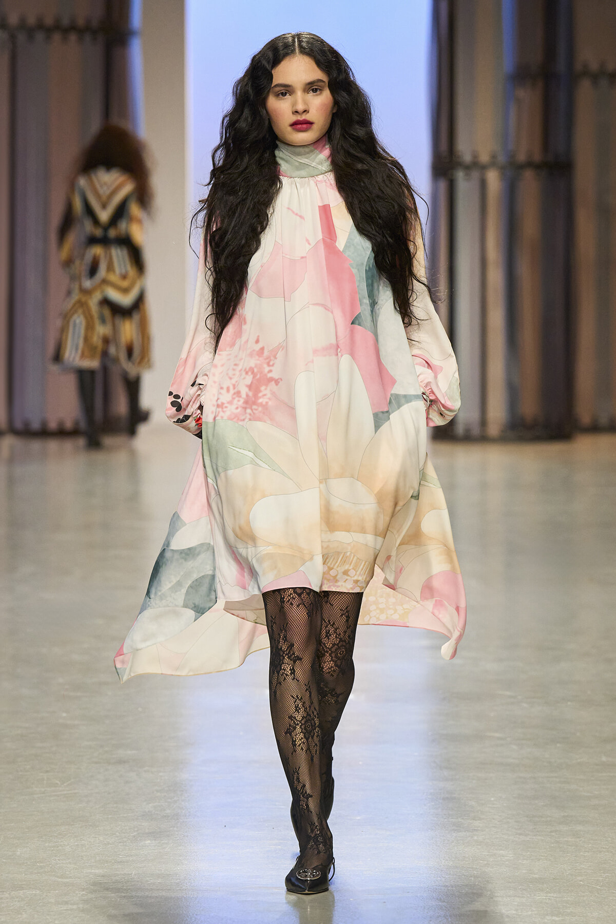 Model walking a fashion show runway in a flowing pastel floral dress with long sleeves and black lace tights.