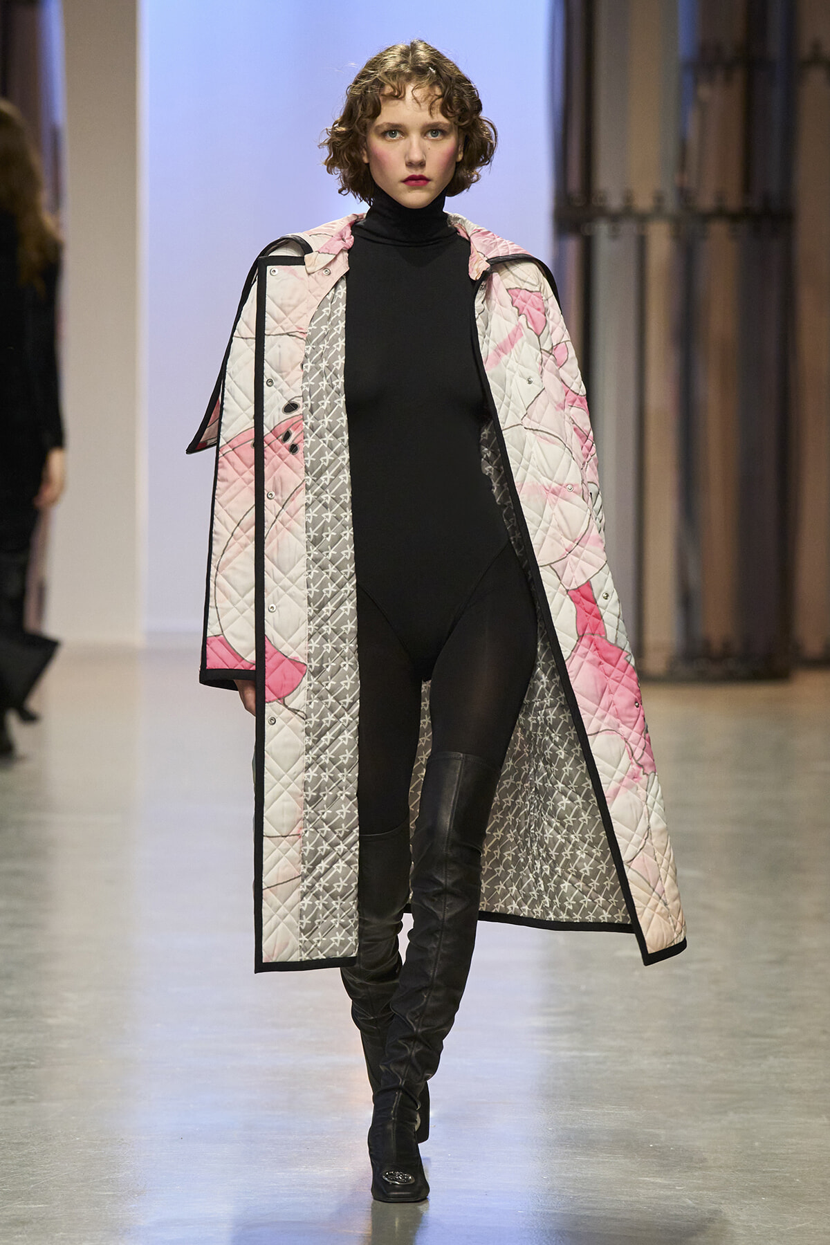 Model walking runway in a long quilted coat with pink, gray, and black patterns over a black turtleneck and black leggings.