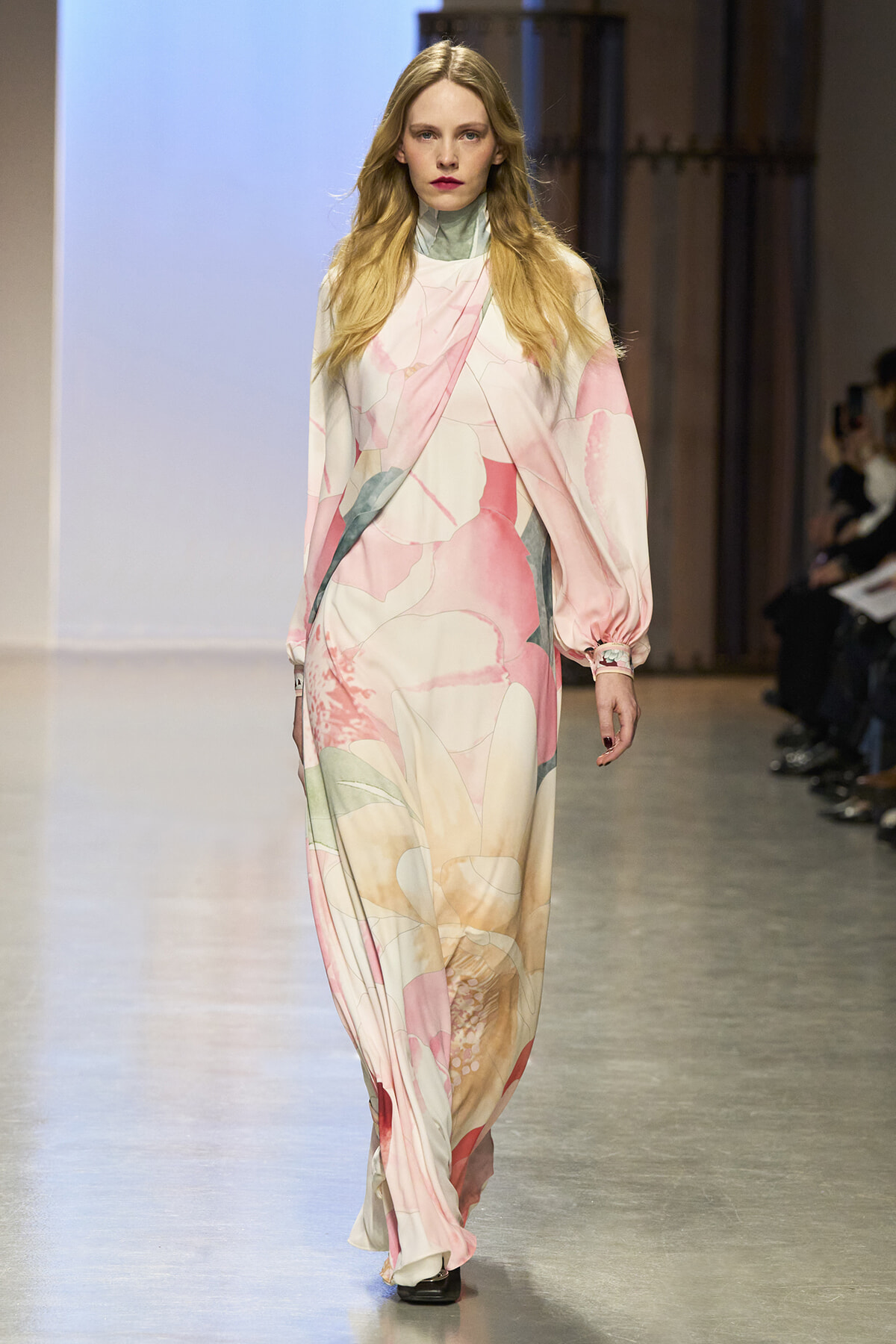 Model walking a fashion show in a flowing pastel pink and cream floral maxi dress with a green-tinted collar at the neckline.