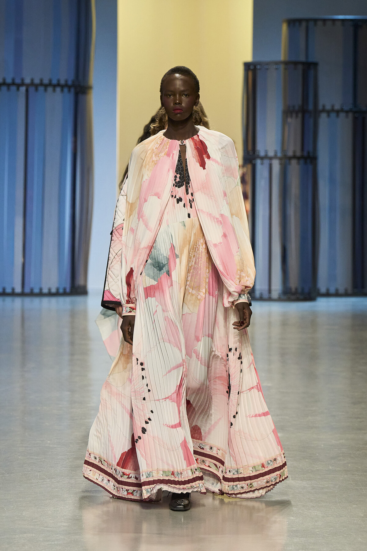 Model walks the runway in a flowing pastel pink dress with abstract brushstroke print and cape-like sleeves on a fashion show catwalk.