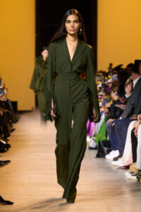 Model walks a runway in an olive-green belted jumpsuit with fringe sleeves and wide lapels, audience in the background.
