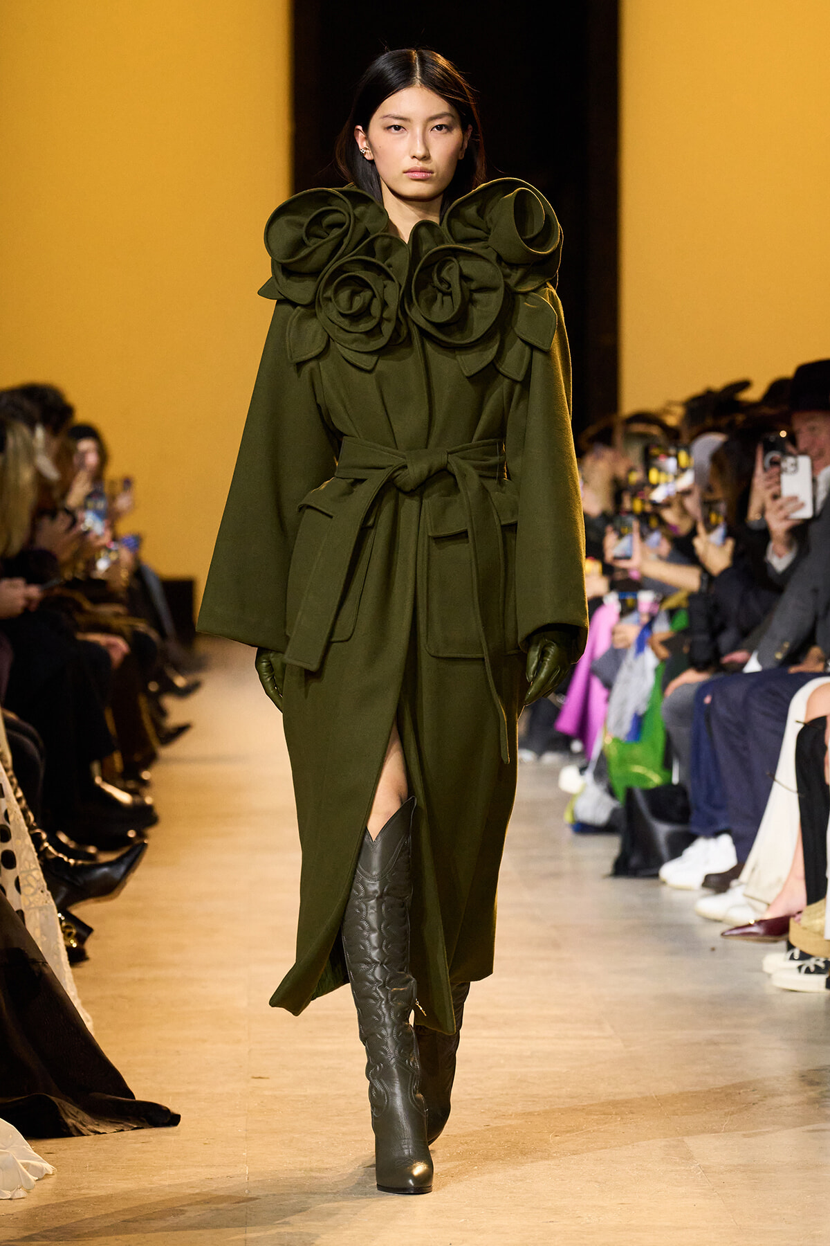 Model on a runway wearing an olive-green coat with oversized rosette shoulders, a tied waist, and knee-high boots.