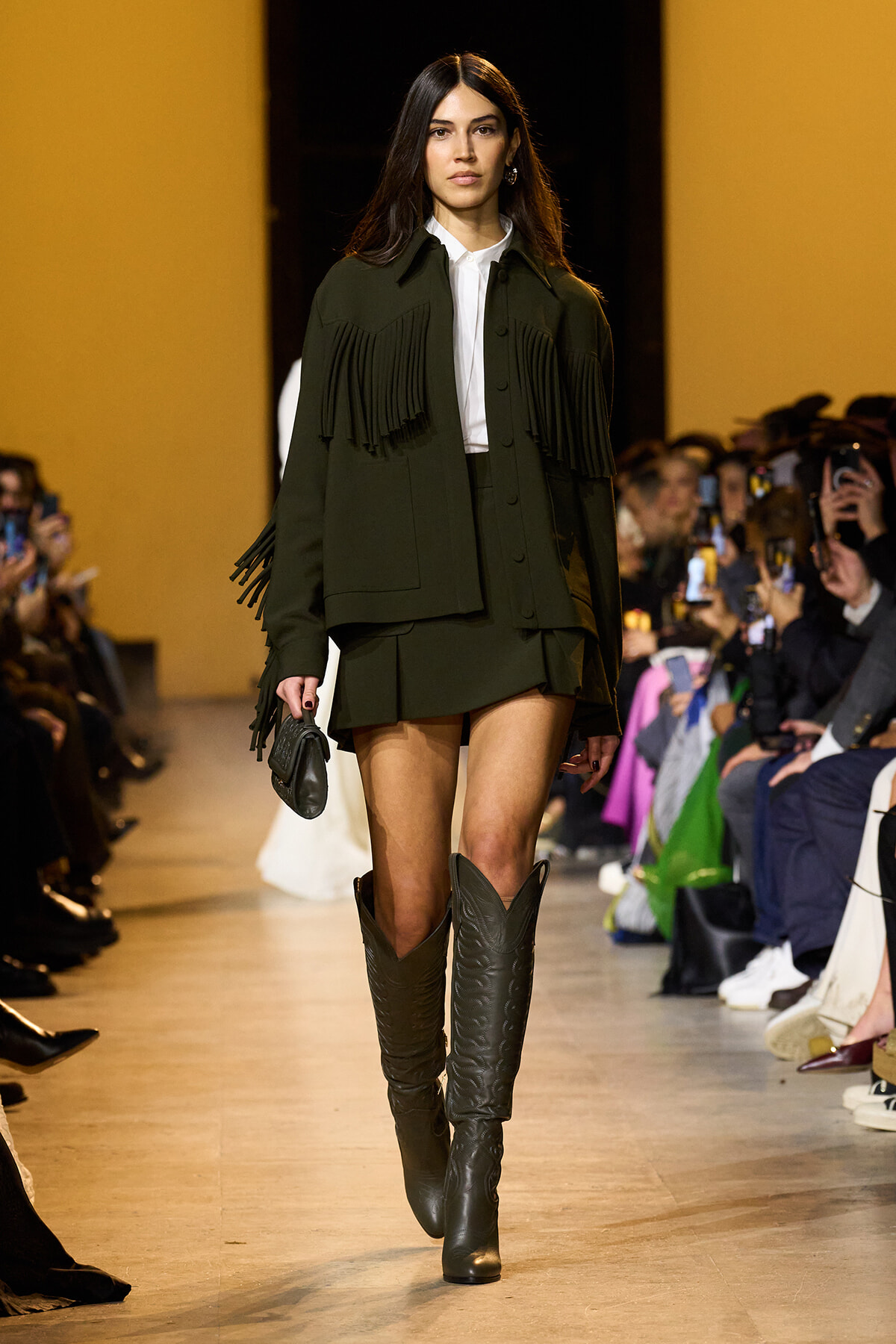 Female model walking a runway in a dark green fringe jacket over a white shirt, matching shorts, and tall black boots, with cheering audience behind.