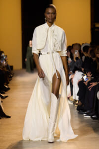 Model walks a fashion runway in an ivory capelet over a white dress with a high slit, paired with tall white boots; audience watches on both sides of the catwalk.