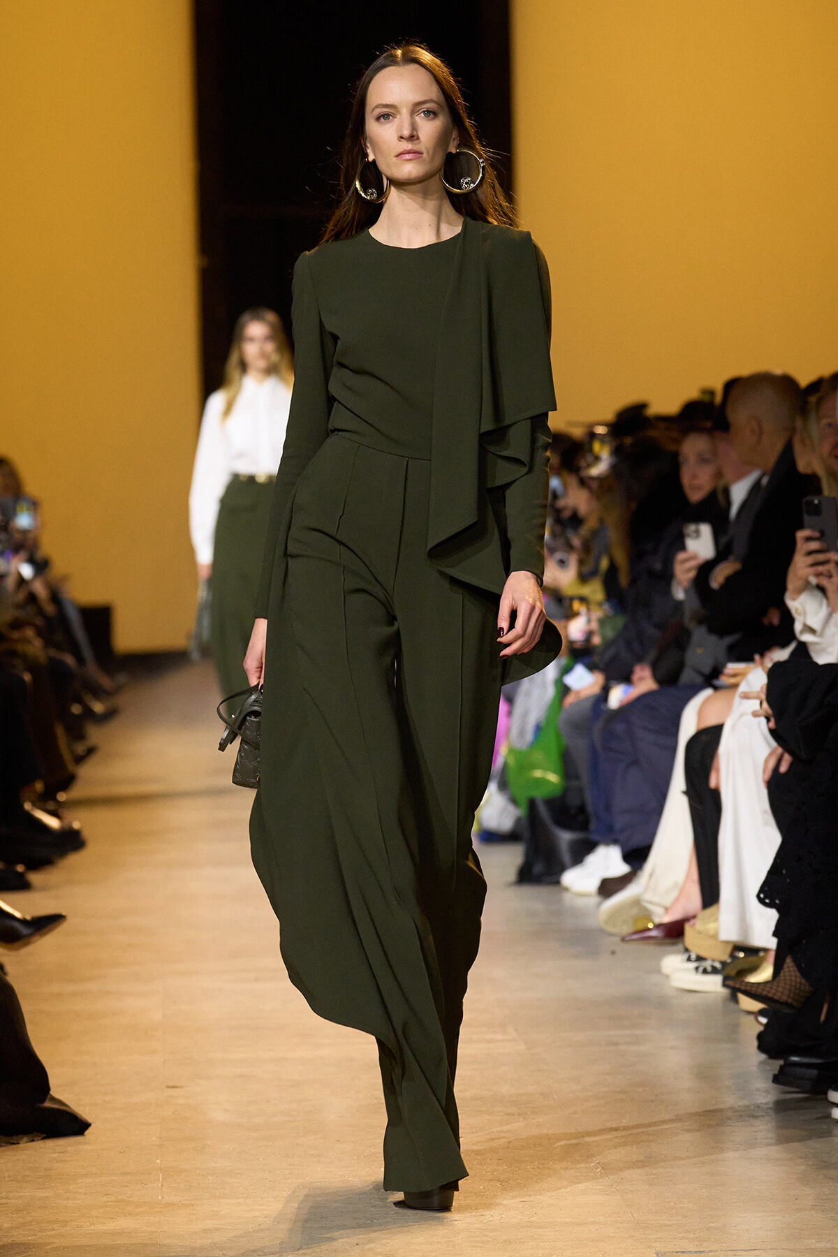 Model walks the runway in a dark olive green draped outfit with oversized sleeves, wearing large hoop earrings and carrying a small black bag; audience lines the catwalk.