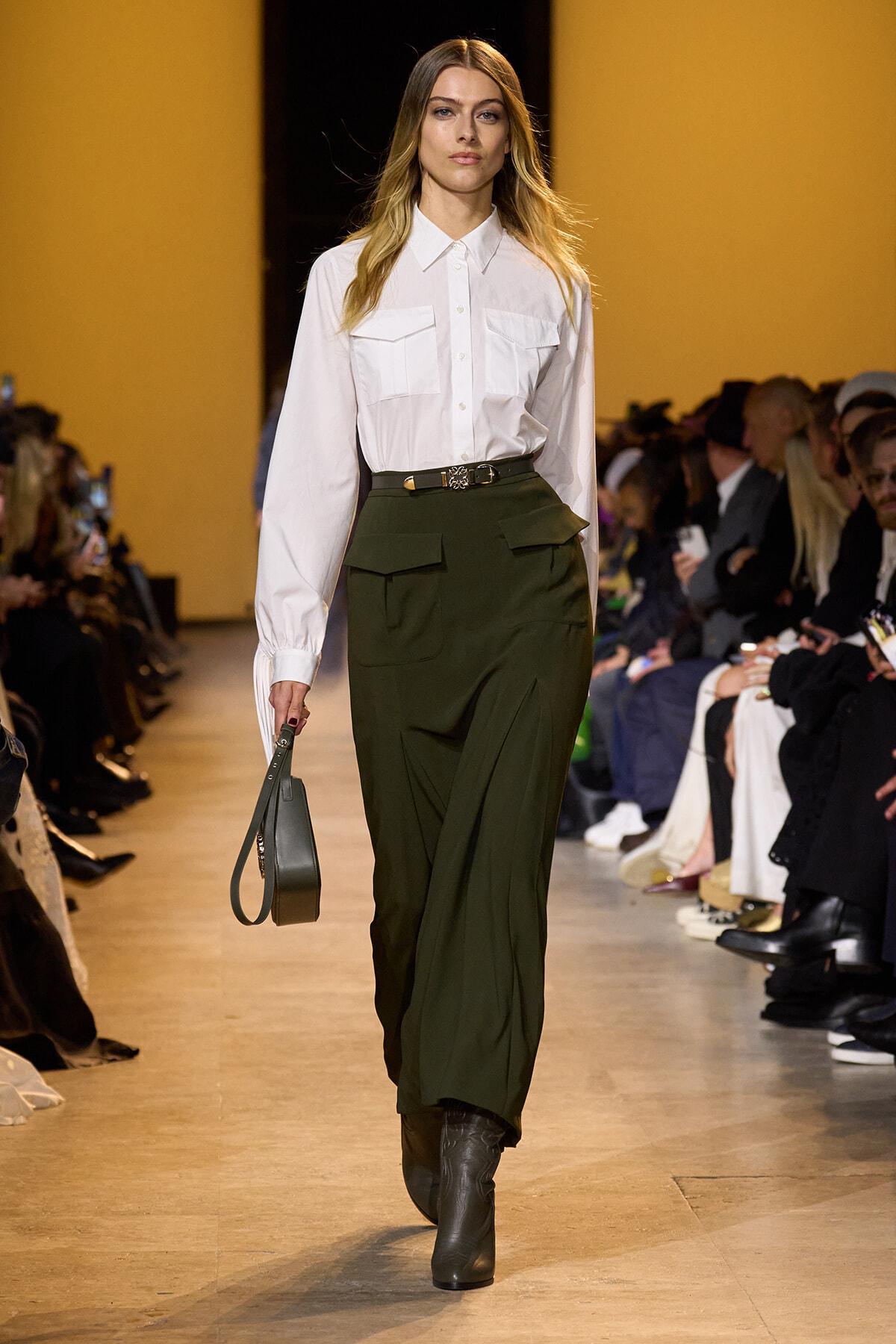 Model walking the runway in a white button-down shirt tucked into olive green cargo pants, with a small gray handbag in hand and boots visible from below.