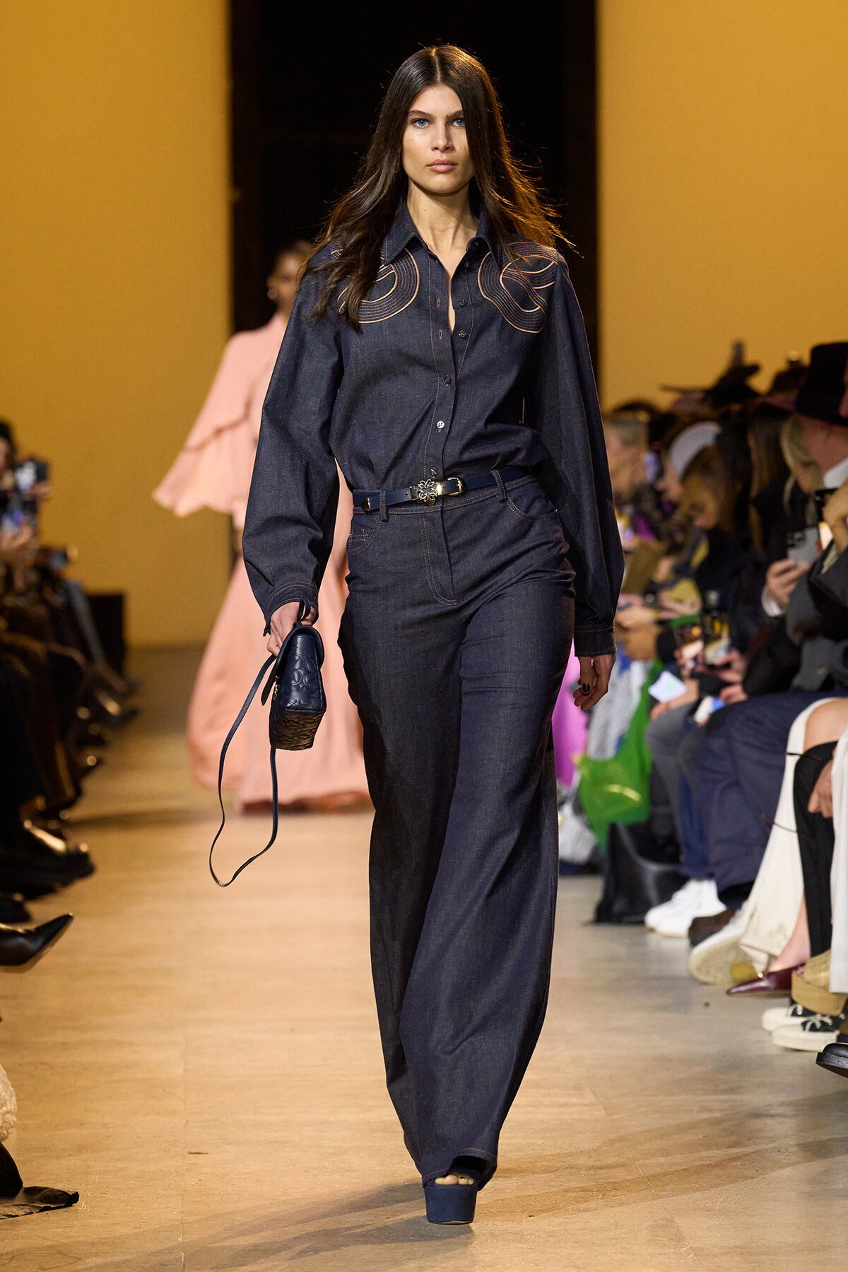 Model walking a runway in a dark denim denim-on-denim outfit with embroidered shoulders, carrying a small navy bag and belt, audience in background