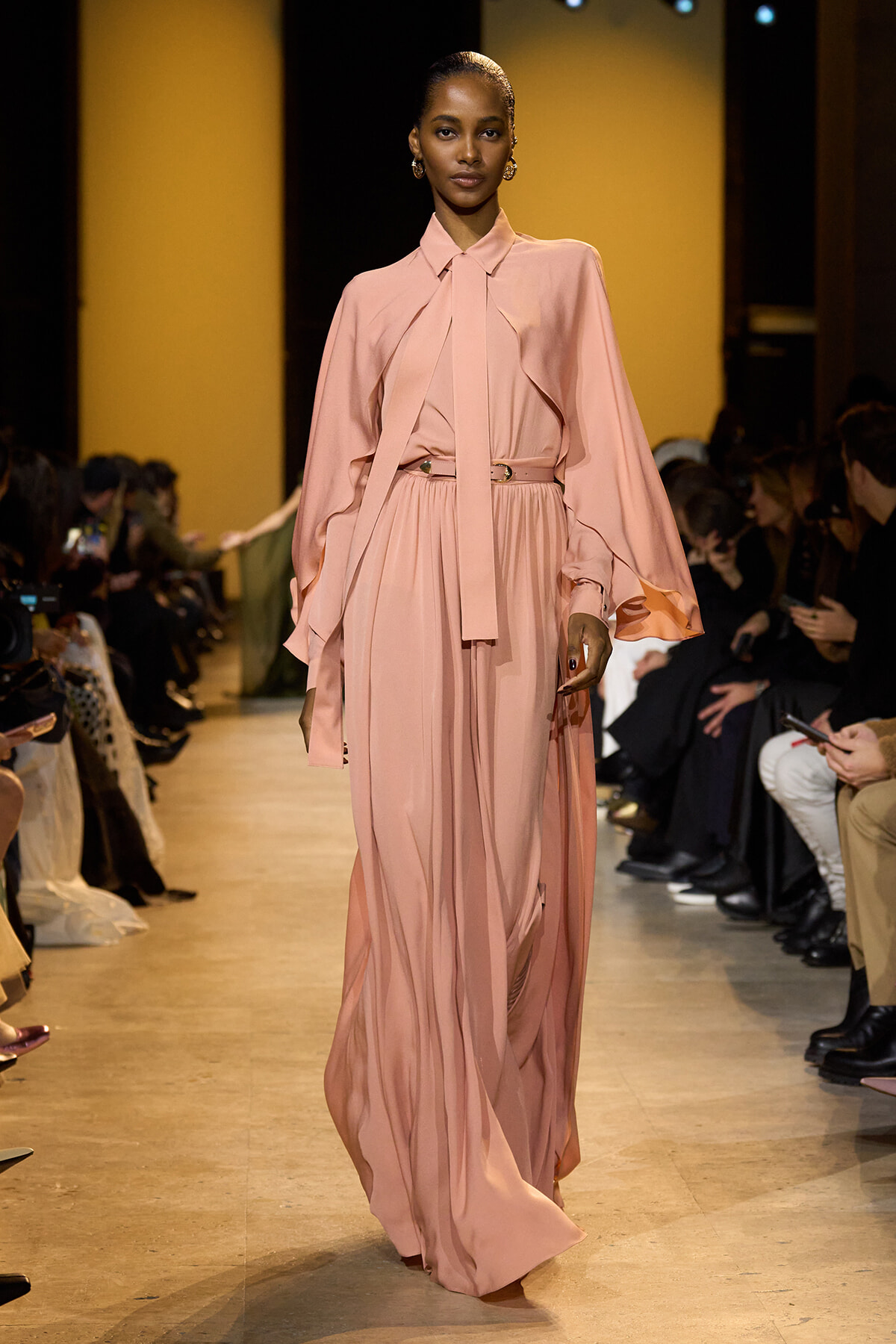 Female model walks a fashion runway wearing a long pale pink dress with cape sleeves and a belt.