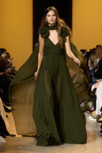 Model walking a runway in a dark emerald green gown with a flowing cape and gathered bodice.