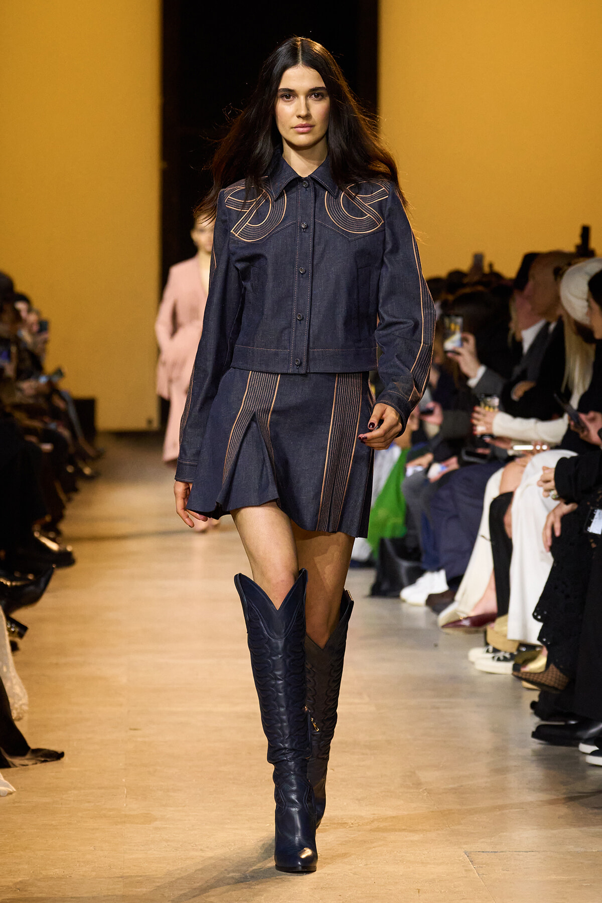 Model walking the runway in a dark denim ensemble with gold-stitched detailing and knee-high blue boots; audience along the sides.