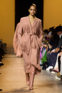 Model on a runway wearing a blush pink fringe coat with a tie belt and matching boots, audience visible on the right.