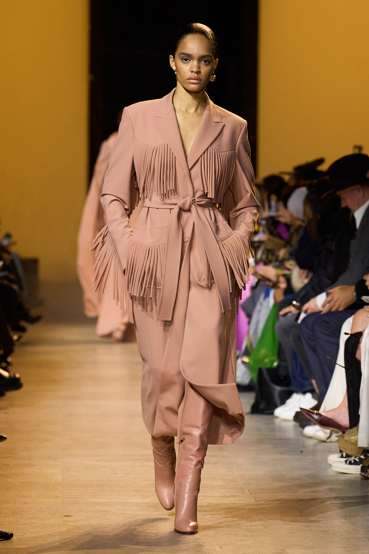 Model on a runway wearing a blush pink fringe coat with a tie belt and matching boots, audience visible on the right.