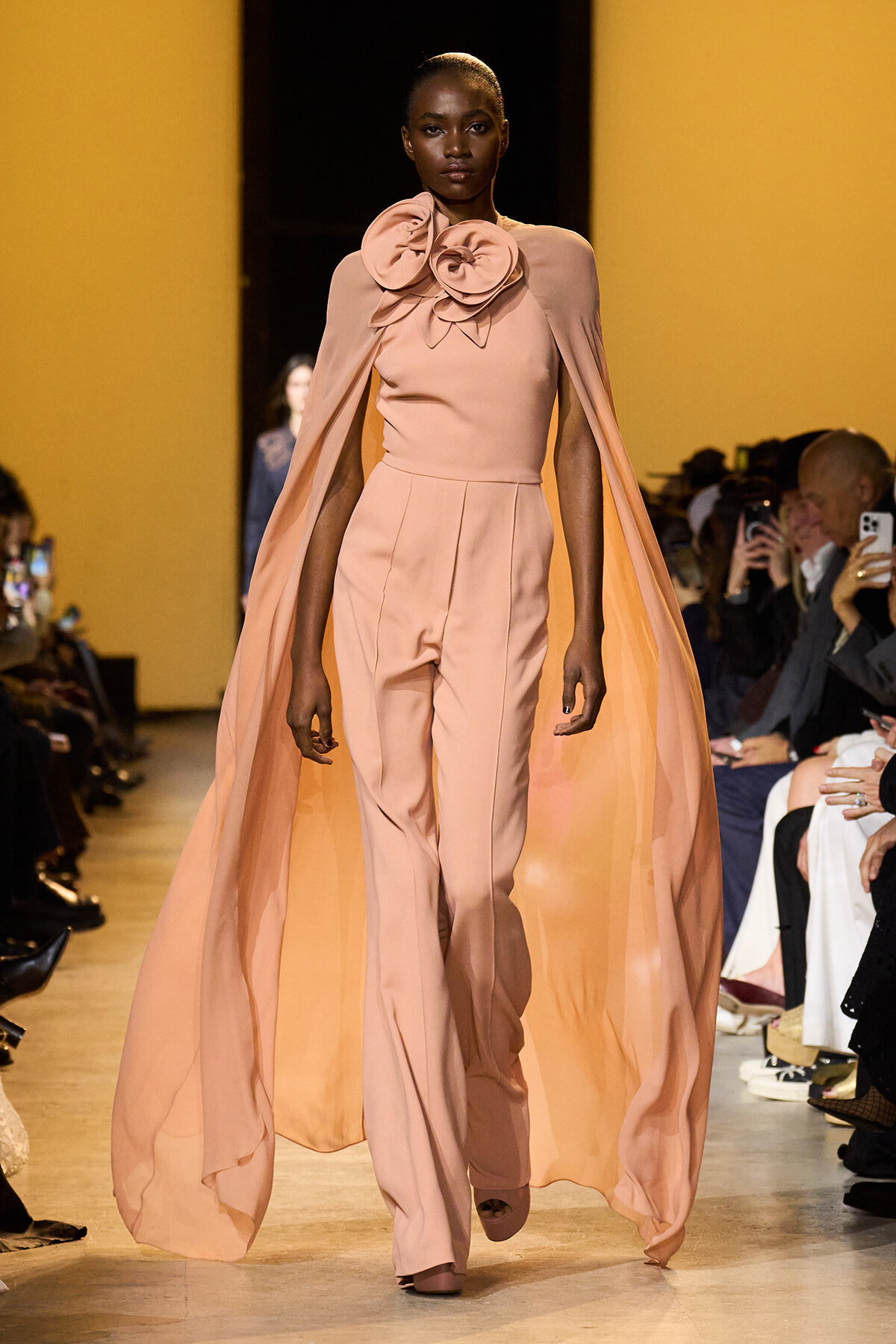Model walking the runway in a peach-pink ensemble: wide-leg pants, fitted top, a dramatic cape, and a large fabric rose at the neck.