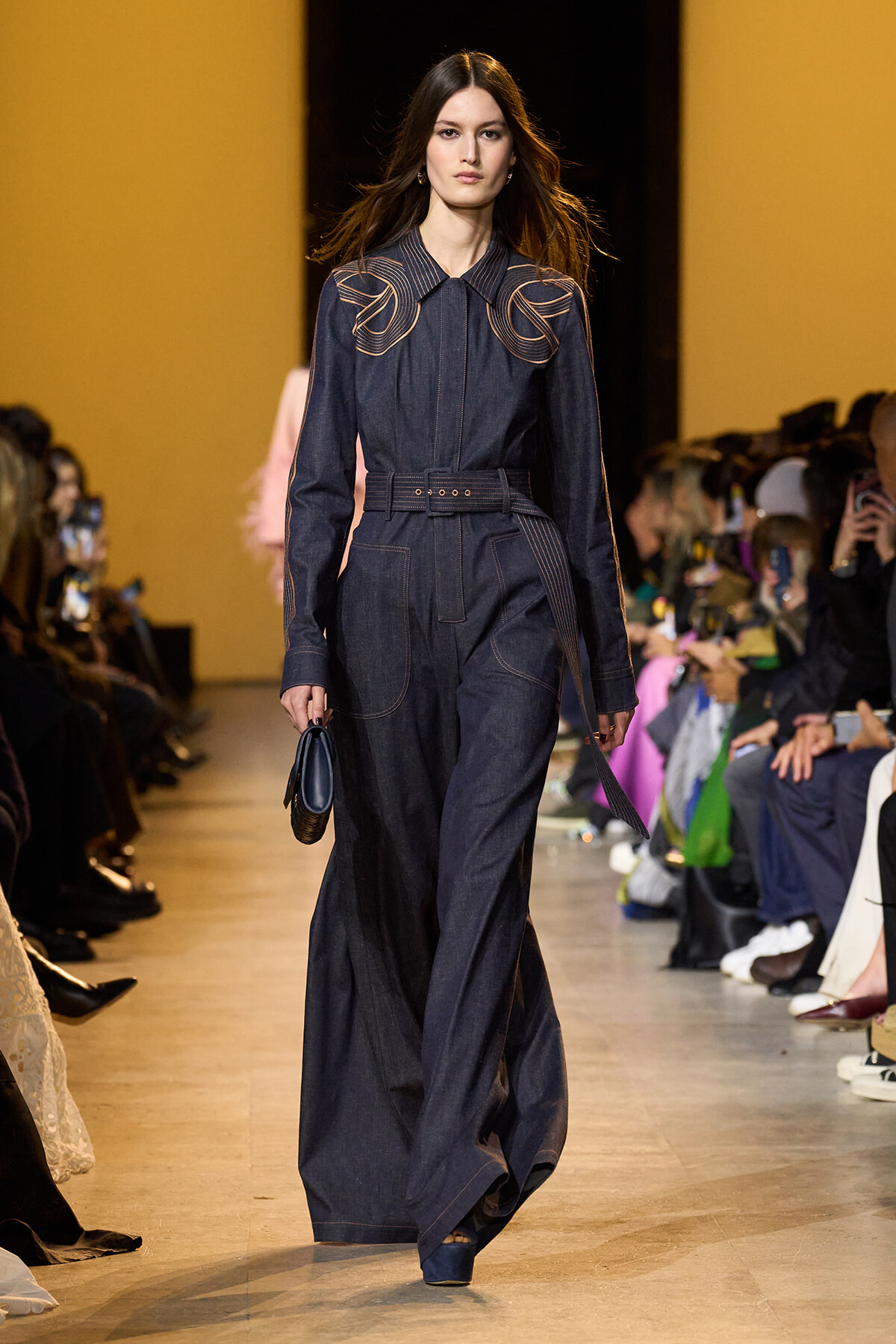 Model walking the runway in a dark denim jumpsuit with tan contrast stitching and a wide belt, audience on both sides taking photos in a fashion show.