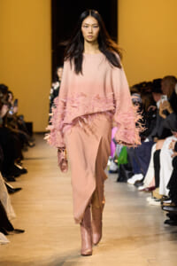 Model walks the runway in a soft pink knit sweater with feathered fringe and matching skirt, pink boots, carrying a small pink handbag.