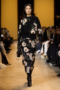 Female model walking a fashion runway in a black floral-knit sweater and matching sequined floral skirt, wearing black gloves with an audience in the background.