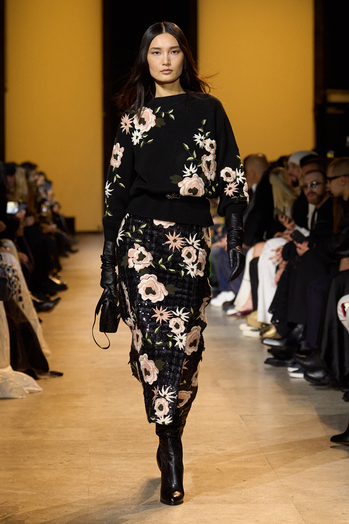 Female model walking a fashion runway in a black floral-knit sweater and matching sequined floral skirt, wearing black gloves with an audience in the background.