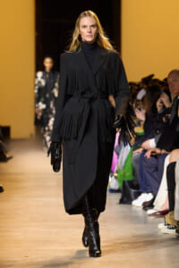 Model on a runway wearing a long black fringe coat with a belt, turtleneck, gloves, and knee-high boots.