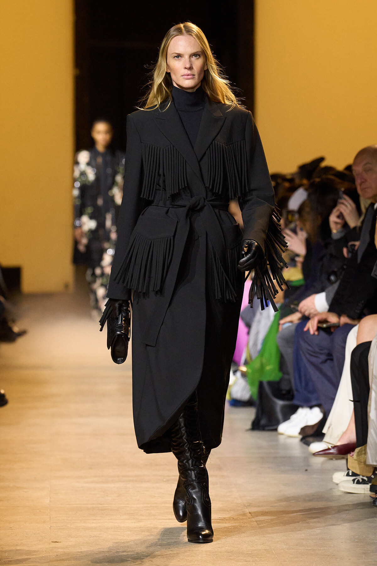 Model on a runway wearing a long black fringe coat with a belt, turtleneck, gloves, and knee-high boots.