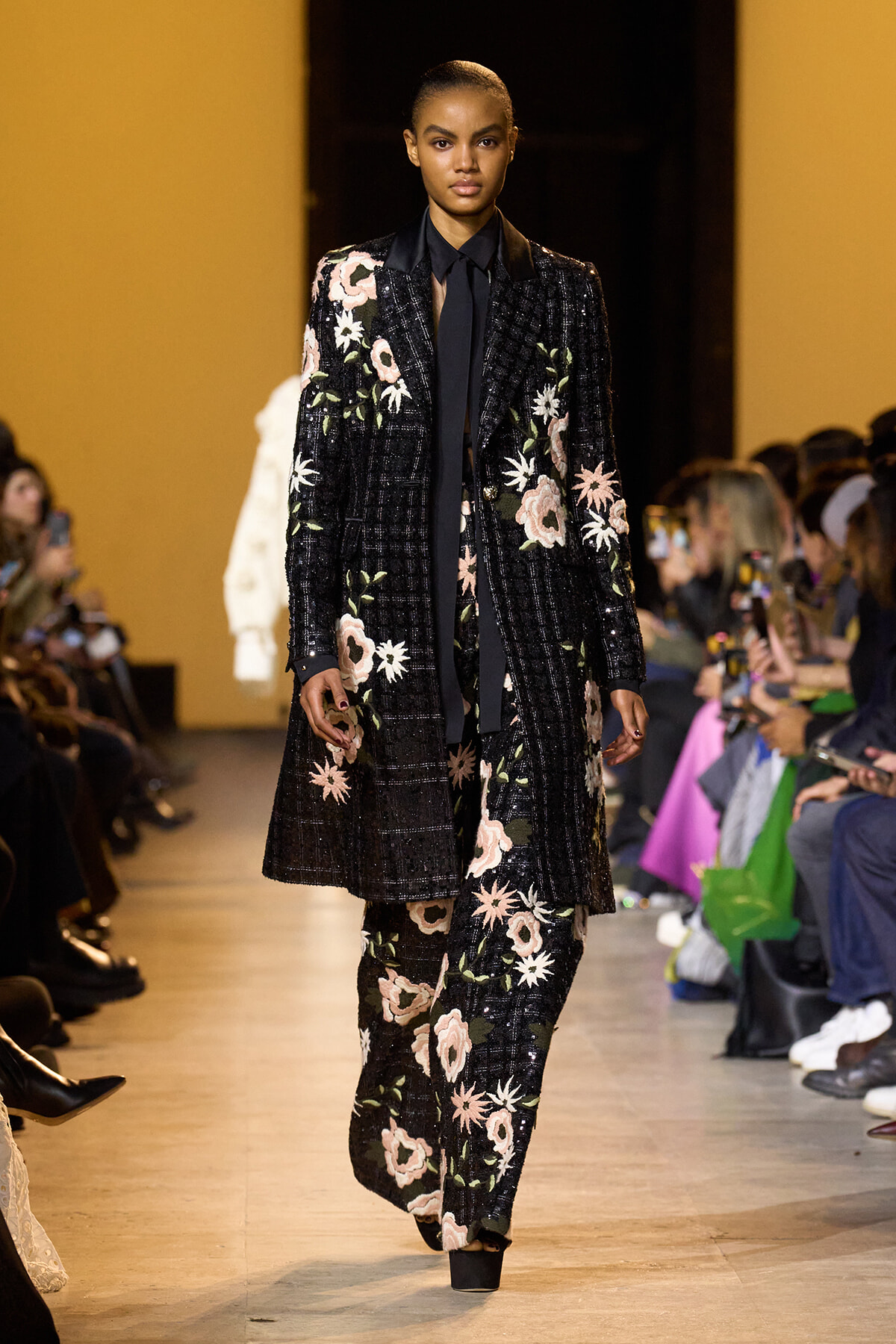 Model on runway wearing a black sequined suit with pink and white floral embroidery and a black necktie.