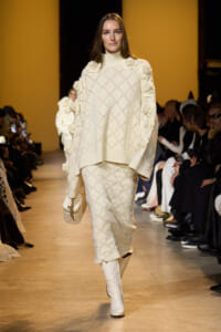 Model walks a runway in an ivory knit sweater and matching skirt with large floral appliqués on the shoulders and sleeves.