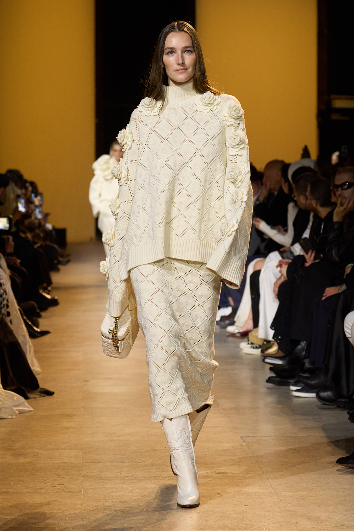 Model walks a runway in an ivory knit sweater and matching skirt with large floral appliqués on the shoulders and sleeves.