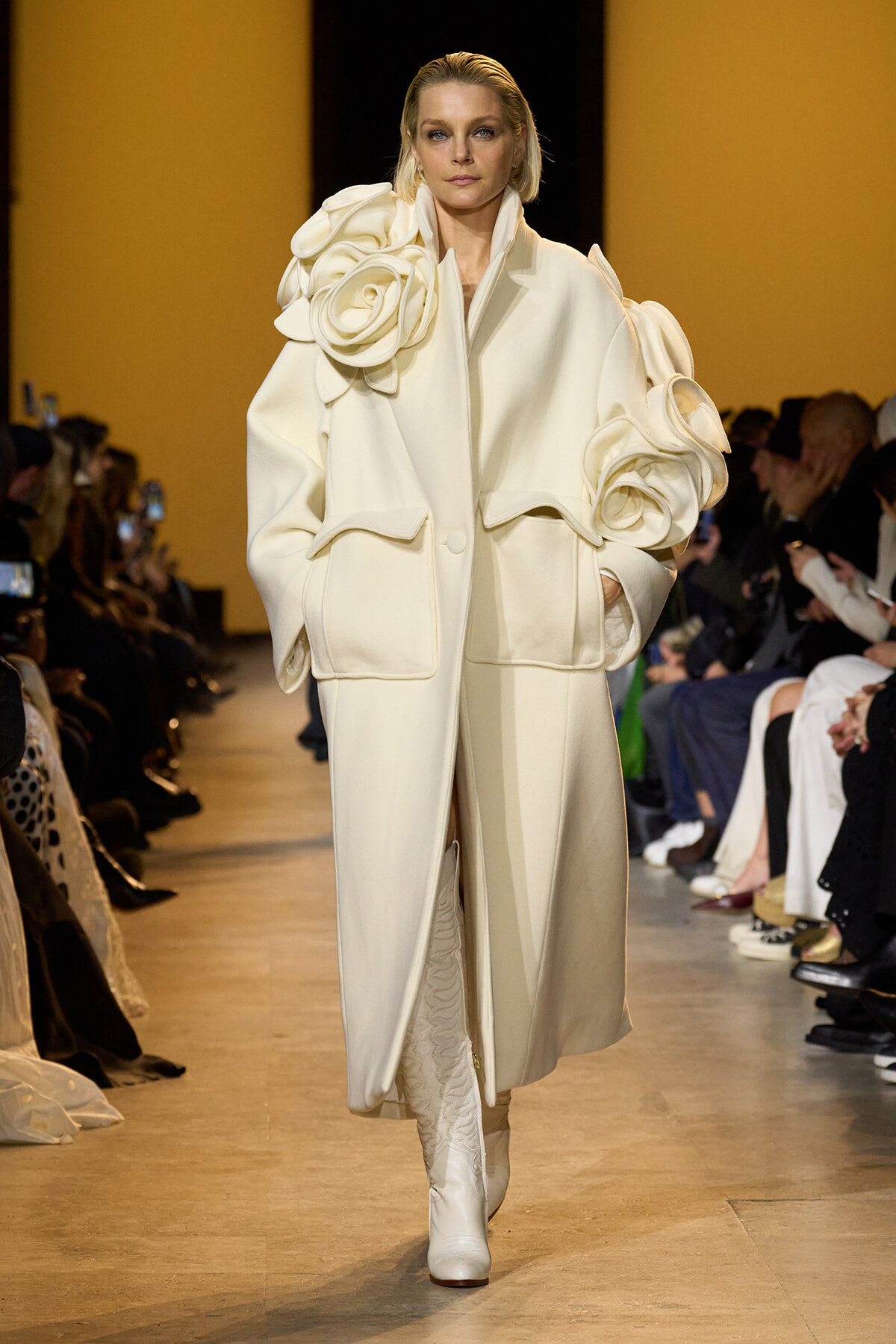 Model walks a runway in an oversized white coat with large fabric rosettes, cream boots, and a pale backdrop.