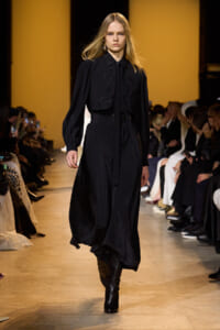Model walking the runway in a black, long-sleeve outfit with decorative piping, audience seated on both sides.