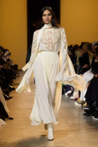 Model walks a fashion runway in a white lace blouse with long sleeves and a flowing ivory skirt; yellow backdrop. (informative)