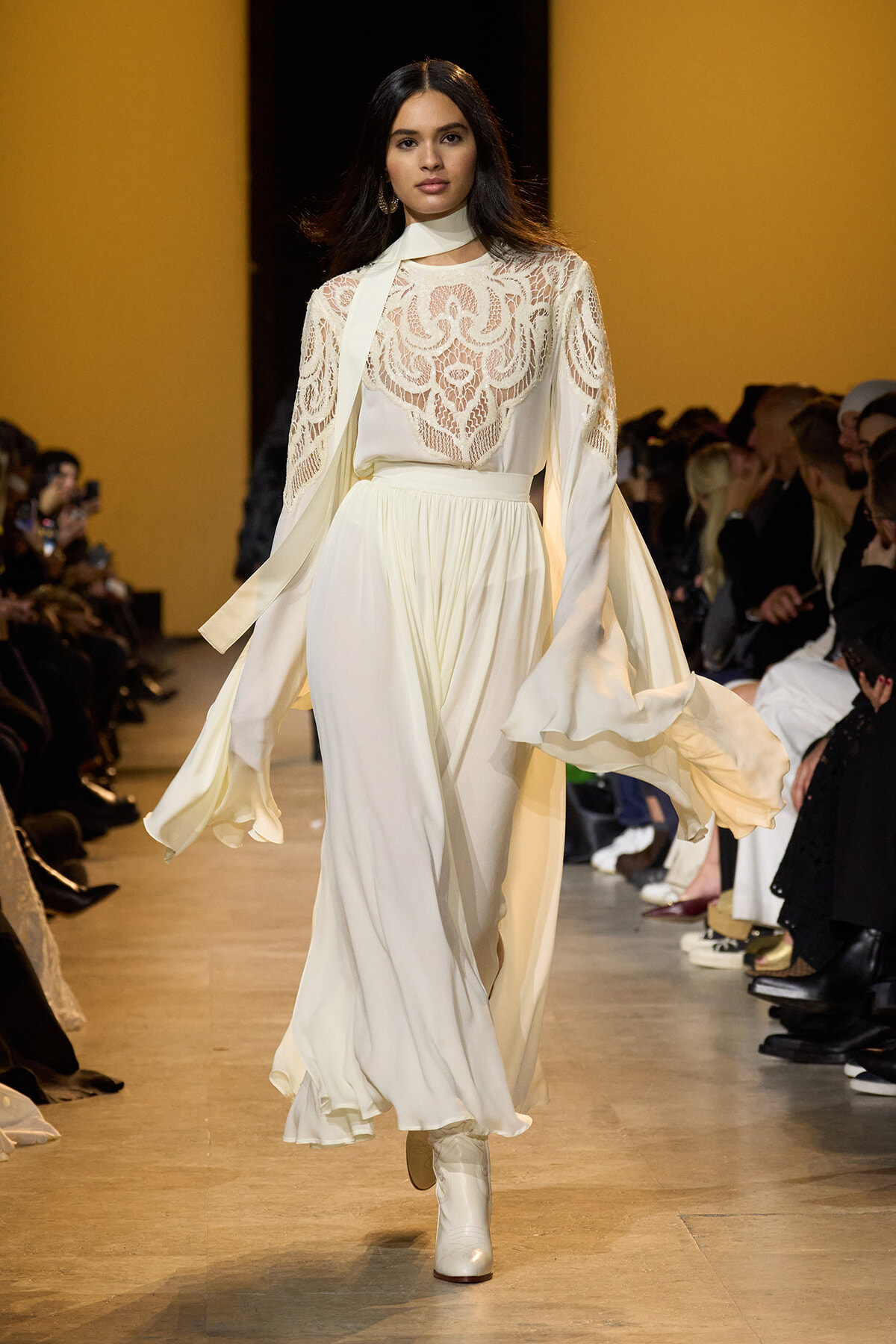 Model walks a fashion runway in a white lace blouse with long sleeves and a flowing ivory skirt; yellow backdrop. (informative)
