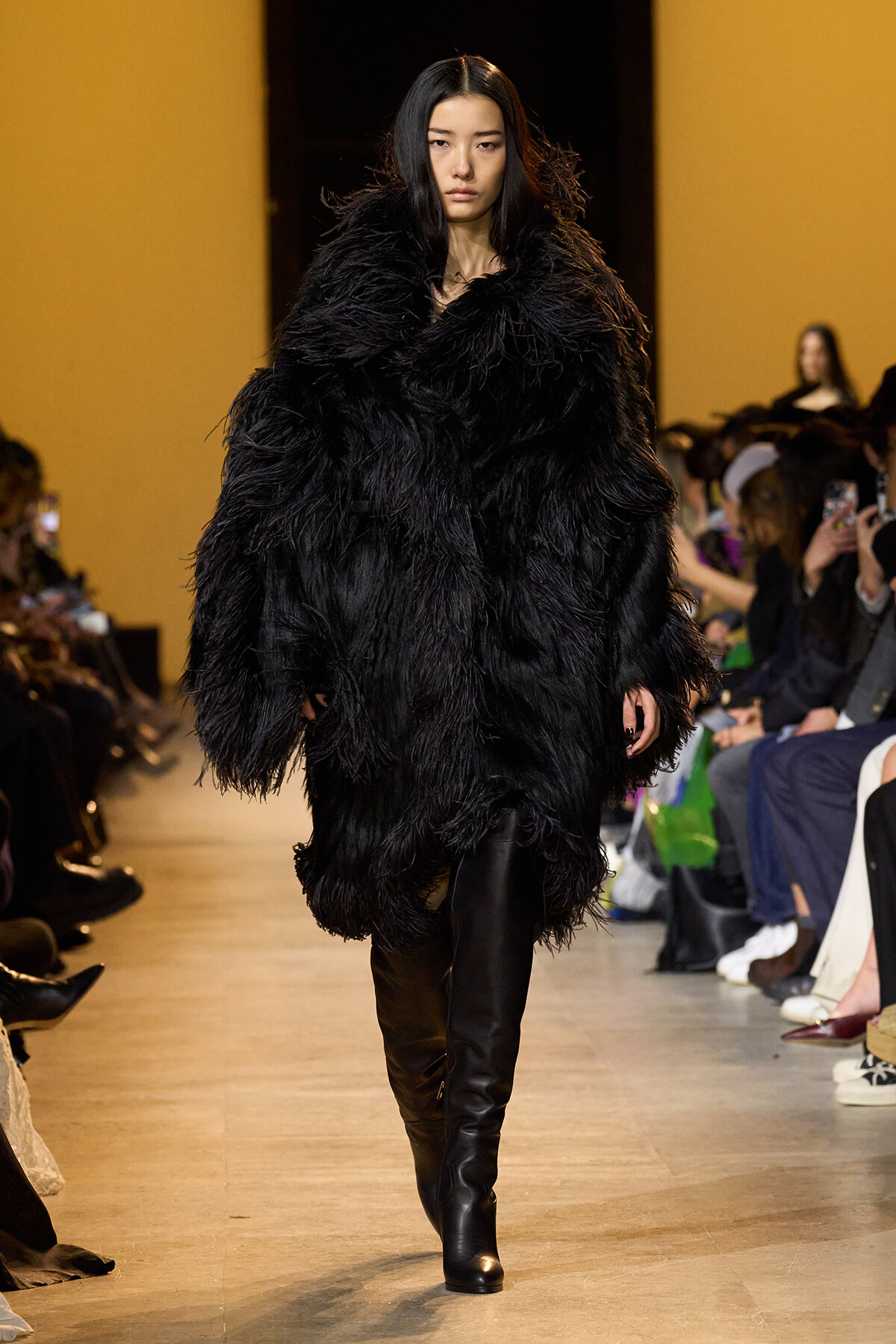 Female model walks a runway in an oversized black feathered coat with knee-high boots, audience blurry in the background