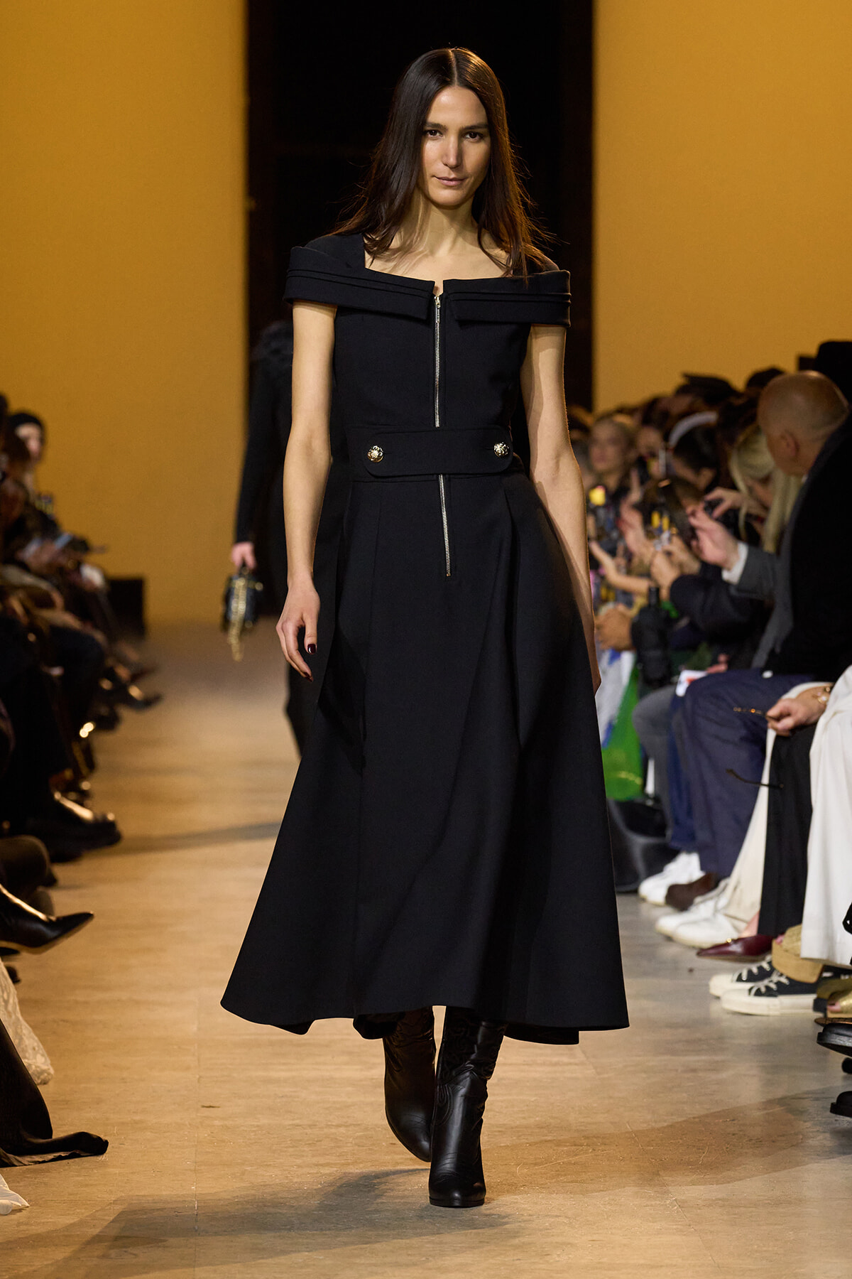 Model walking a fashion show runway in a black off-the-shoulder dress with a front zipper and wide jeweled belt, black boots, and an audience in the background