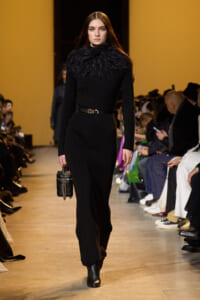 Model on a runway wearing a black knit dress with a voluminous feather collar, belted at the waist, carrying a small black handbag.
