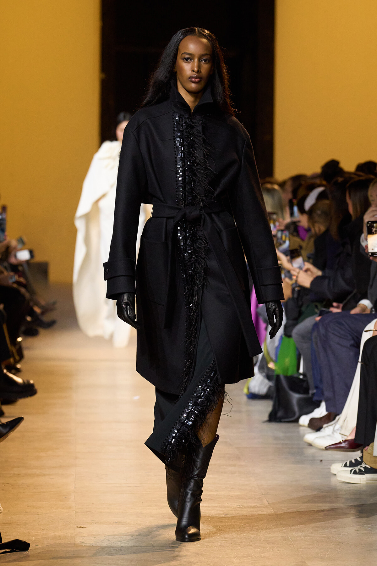Female model walking a runway in a black knee-length coat with fringe detailing, belted at the waist, paired with black gloves and boots.