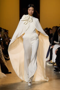 Model walks the runway in a white cape and matching pants with a large fabric rose embellishment at the shoulder.
