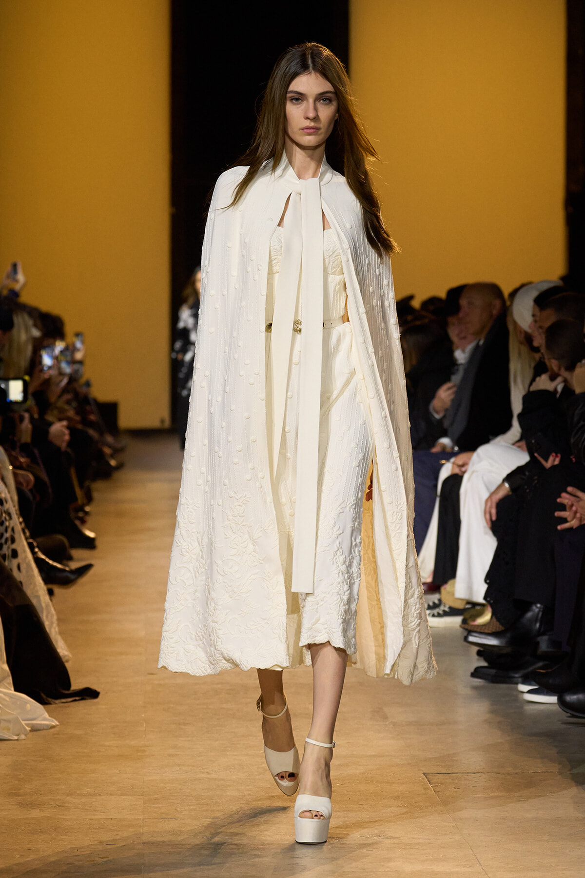 Model on a runway wearing a white embroidered coat and matching dress, beige backdrop behind her, audience on sides